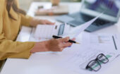 Person reviewing financial documents with a pen, calculator, and glasses on a desk, with a laptop and charts in the background.