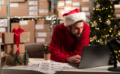 A man in a red sweater and Santa hat works on a laptop at a desk with shipping boxes and labels, with a Christmas tree in the background.