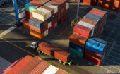 Aerial view of stacked shipping containers and a truck loaded with a container at a cargo terminal.