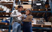 Two warehouse workers standing in an aisle, one using a tablet and the other holding a clipboard.