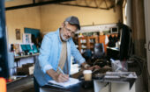 A man in a cap and denim shirt taking notes in a notebook at a desk in a workshop.
