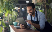 Man wearing an apron smiles while using a tablet at a counter surrounded by green plants.
